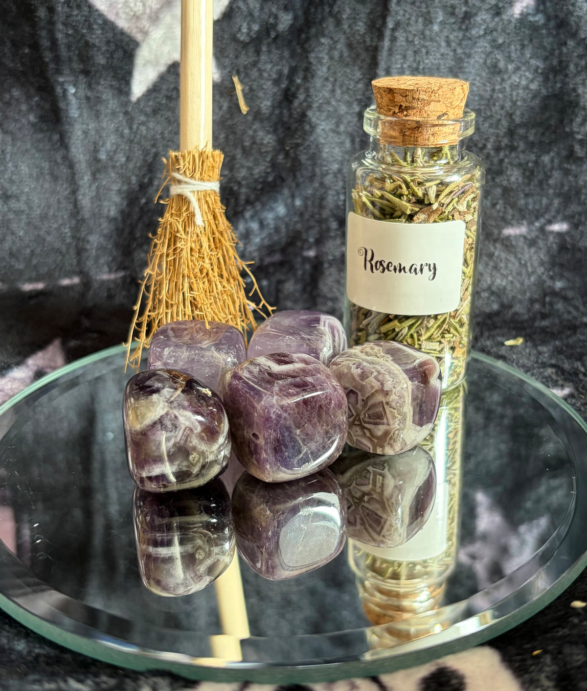 A glass jar labeled 'Rosemary' filled with dried herbs sits on a round mirrored tray beside polished purple stones and a small straw broom against a dark textured backdrop.