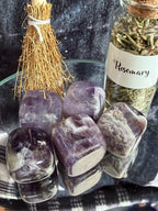 A set of polished purple and white amethyst tumbled stones arranged on a reflective glass surface, next to a small dried rosemary jar and a small straw broom, on a dark textured background.