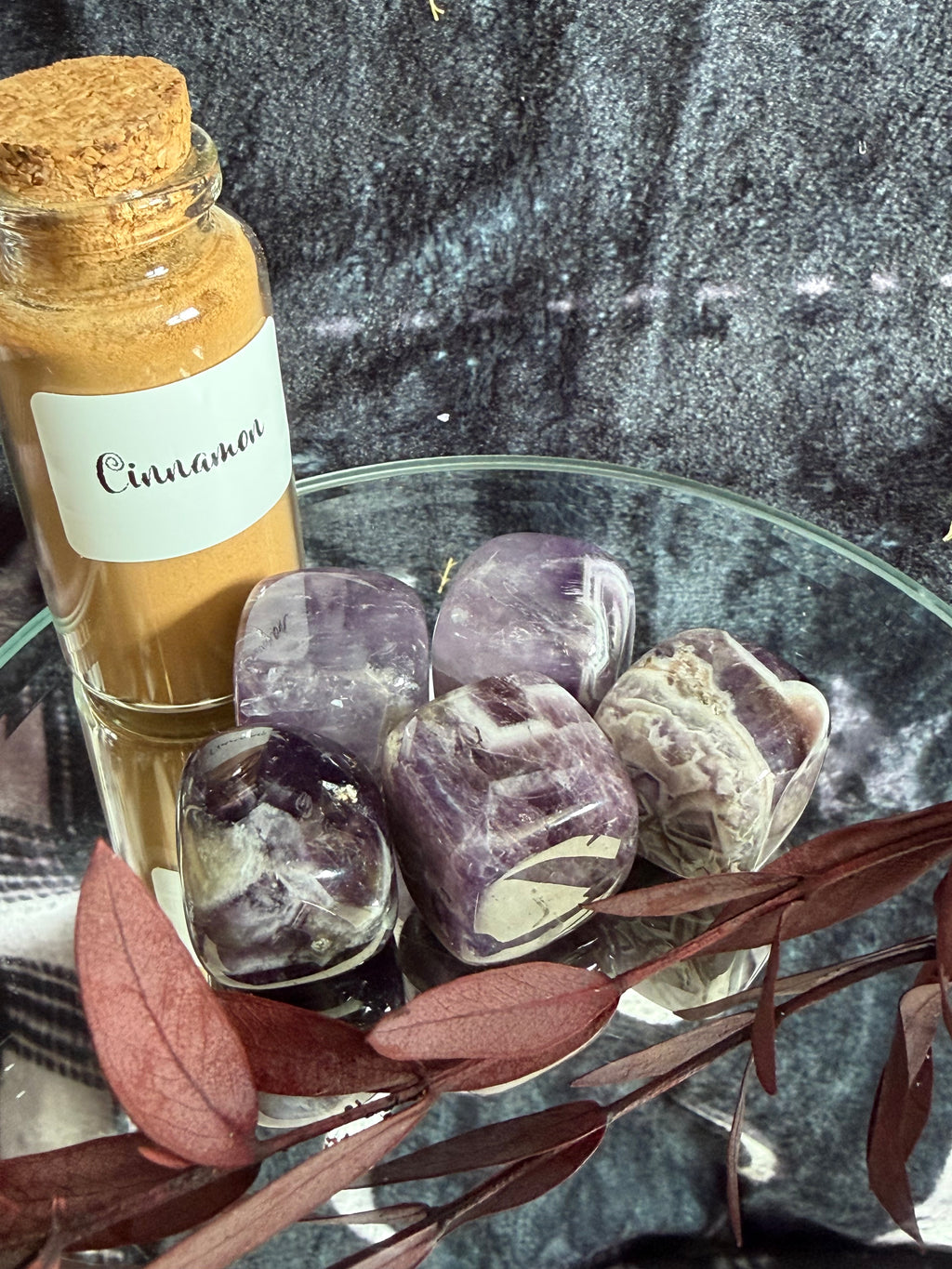 A small glass bottle with a cork stopper and a white label reading 'Cinnamon' filled with warm orange-brown powder, styled beside polished purple crystal stones on a glass plate with dried leaves against a dark textured backdrop.