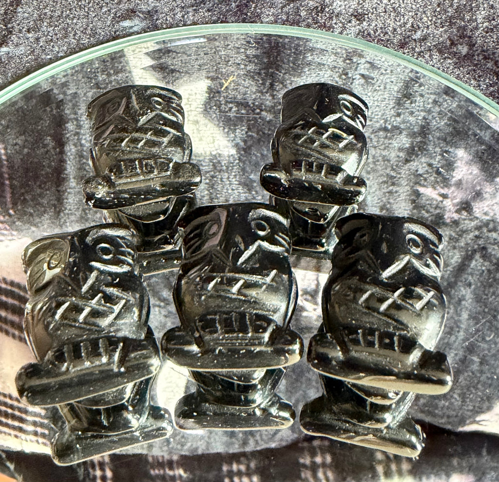 Five small black carved figurines with stylized faces and geometric details, arranged on a clear glass plate over a gray textured surface.