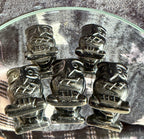 Five small black carved figurines with stylized faces and geometric details, arranged on a clear glass plate over a gray textured surface.