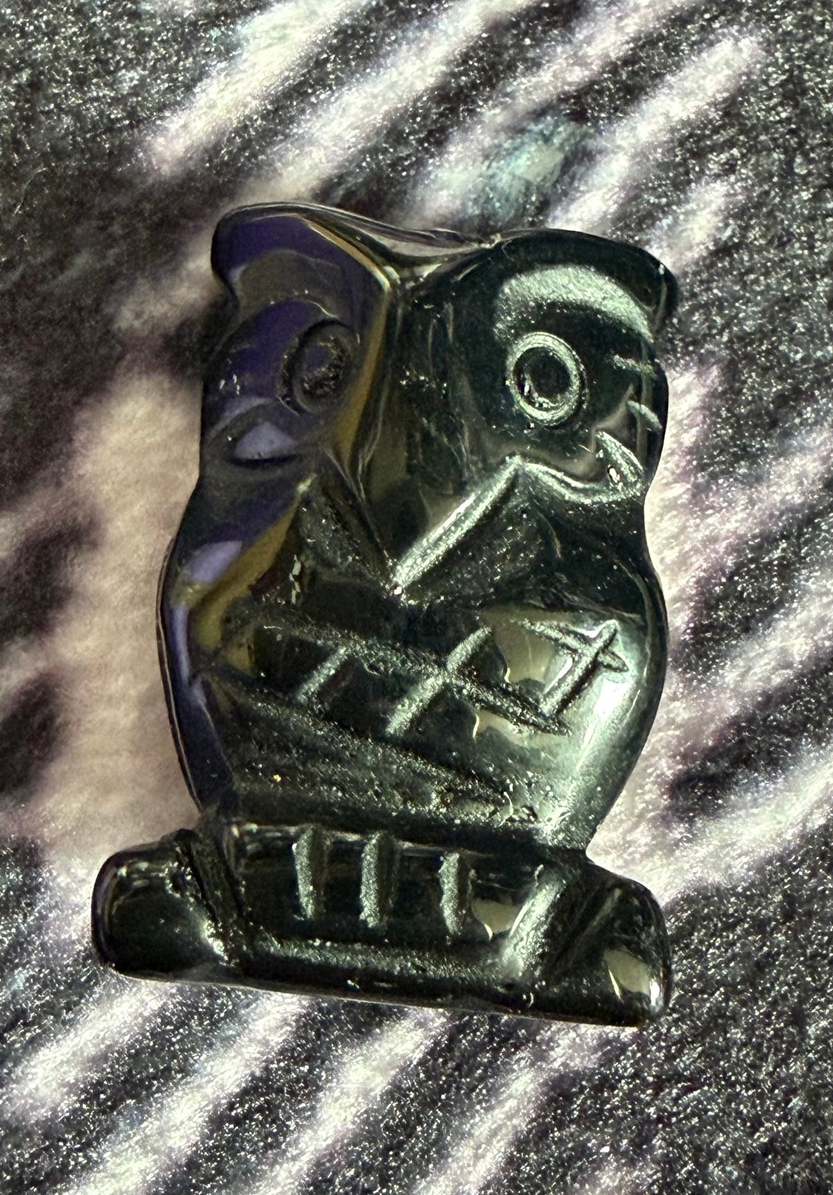 A carved owl-shaped pendant or charm made of dark polished stone, photographed close-up against a sparkly silver and black textured background.