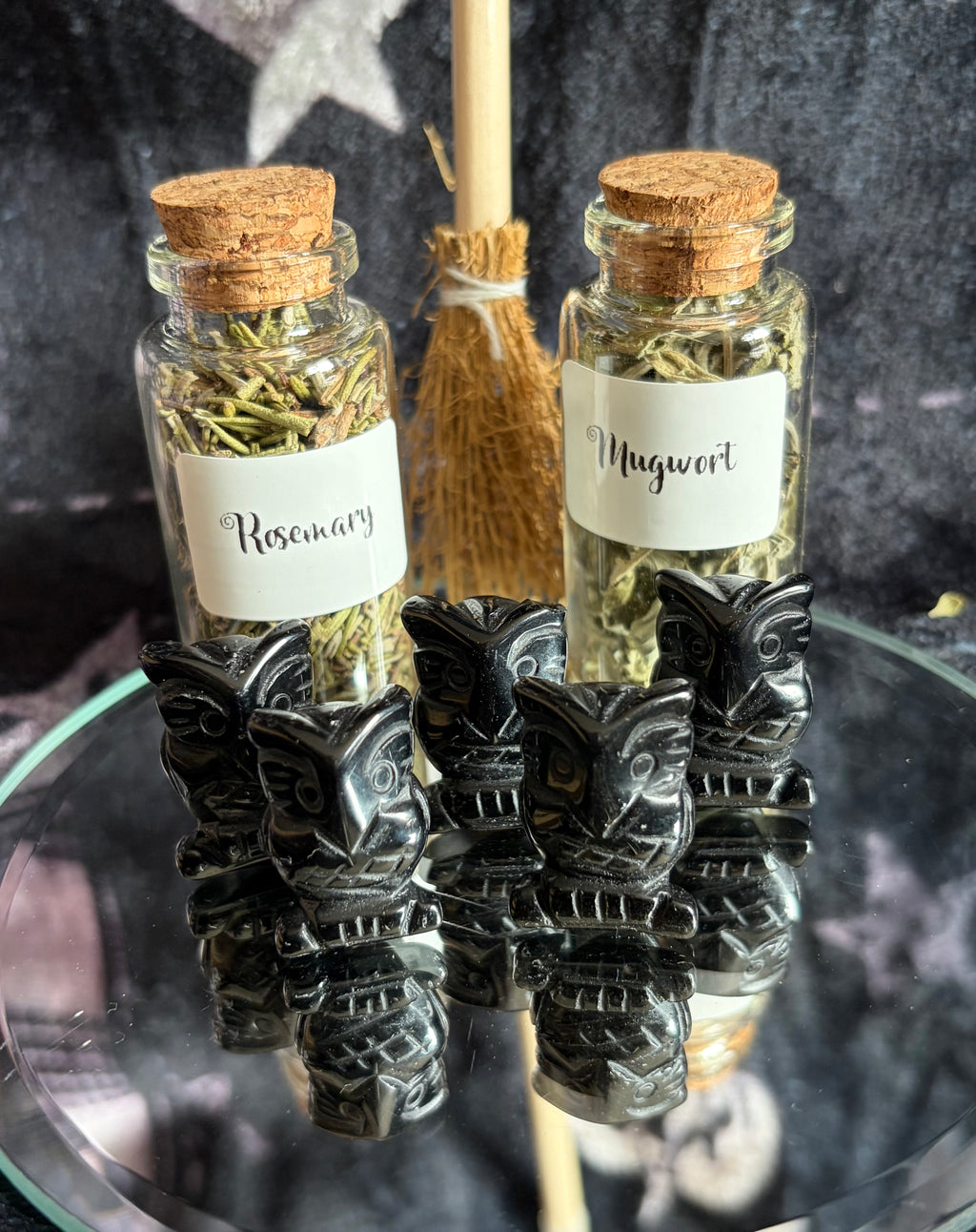 Two small glass vials with cork stoppers, labeled 'Rosemary' and 'Mugwort' with dried herbs inside, displayed on a glass surface with four black carved tiki figurines in front and a small broom behind them.