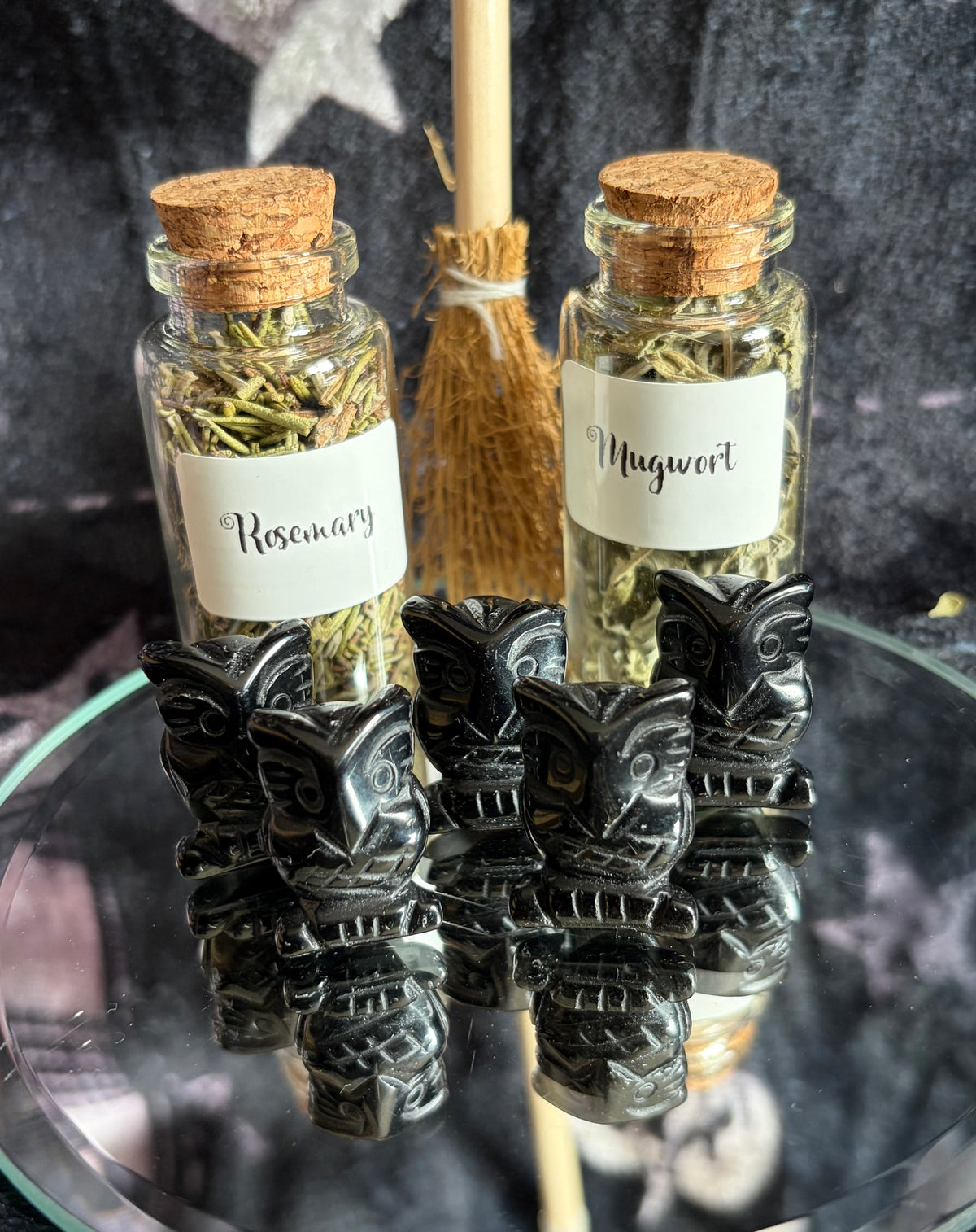 Two small glass vials with cork stoppers, labeled 'Rosemary' and 'Mugwort' with dried herbs inside, displayed on a glass surface with four black carved tiki figurines in front and a small broom behind them.