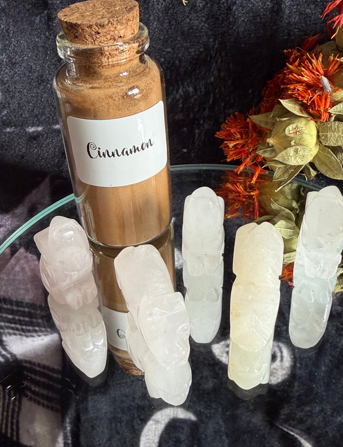 A small glass bottle with a cork stopper labeled 'Cinnamon' filled with brown powder, arranged on a glass surface beside five pale carved crystal points, with dried orange flowers in the background.