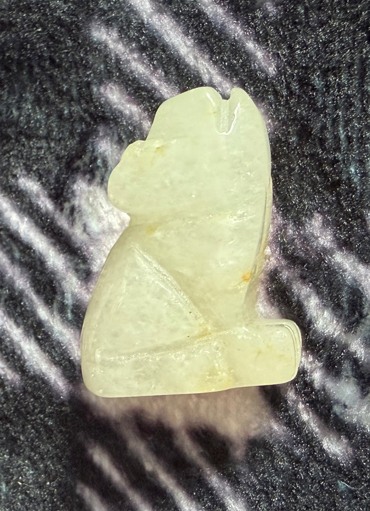A small carved pale yellow-green stone figurine, shaped like a seated animal, photographed on a dark textured surface with bright light reflections.