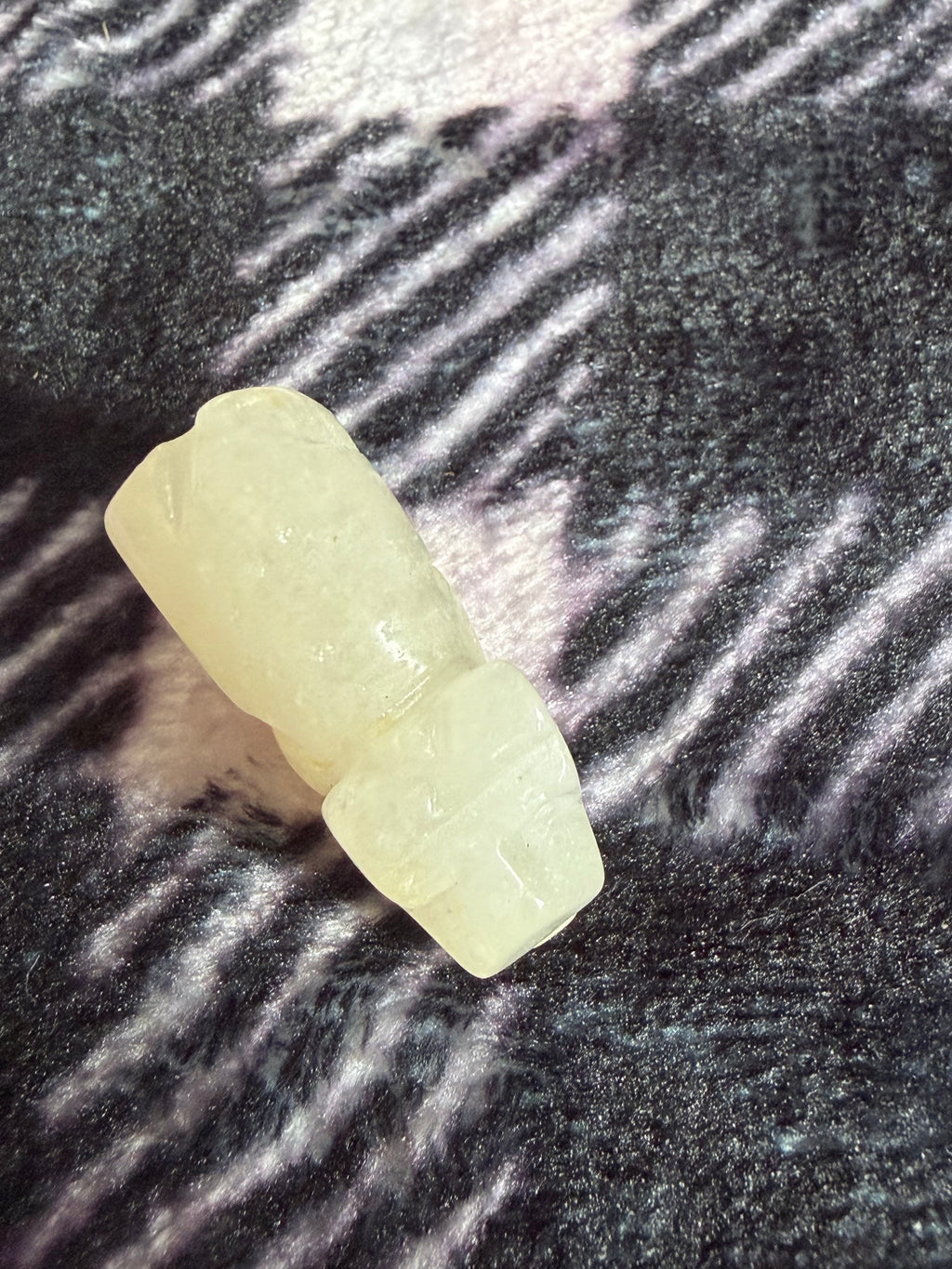 A small pale yellow carved stone or resin figurine placed on a dark textured fabric background with diagonal light reflections.