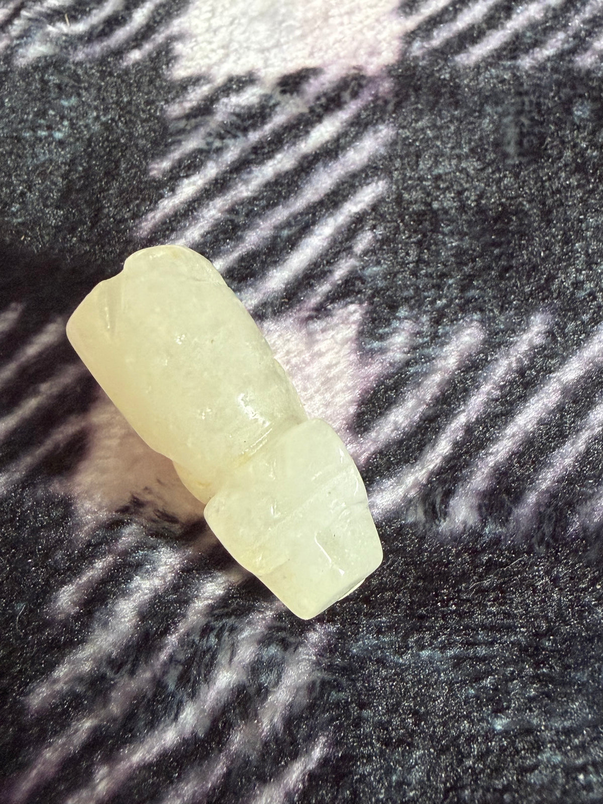 A small pale yellow carved stone or resin figurine placed on a dark textured fabric background with diagonal light reflections.