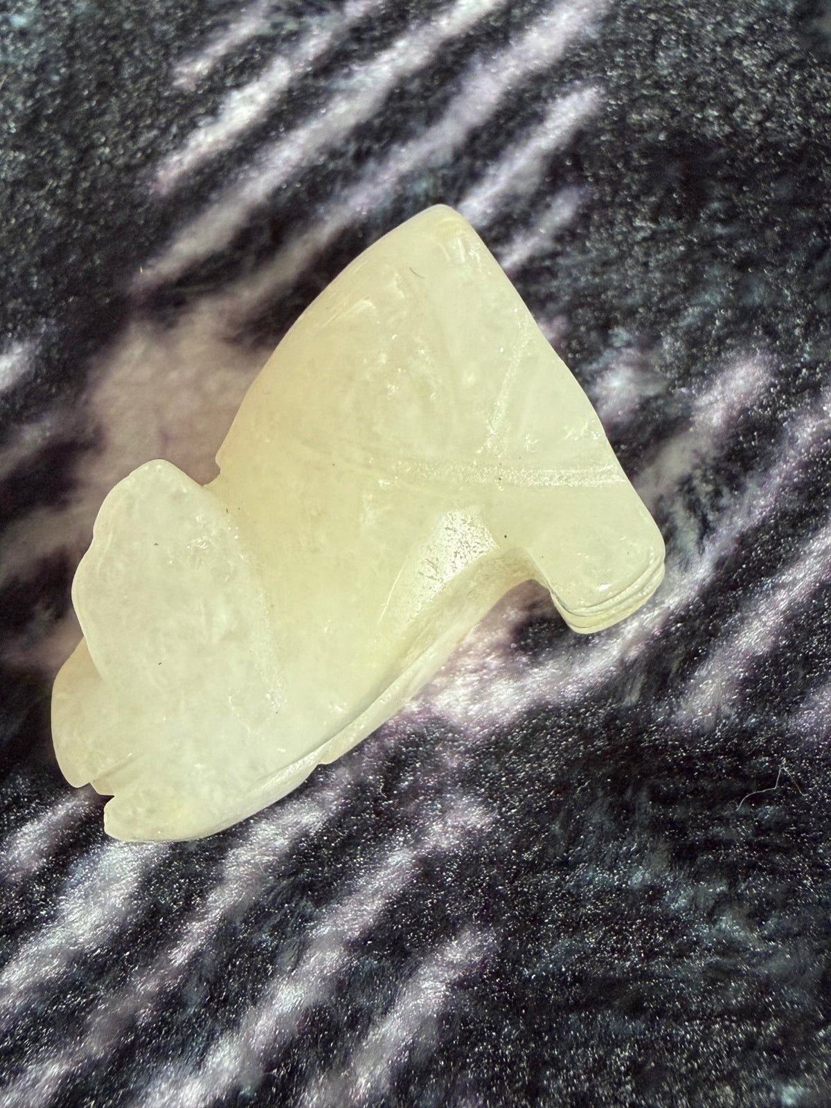 A small carved pale yellow stone or resin piece with a smooth organic shape, photographed on a dark textured surface with bright light reflections.