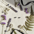 A styled flat-lay of pressed botanical elements on a textured off-white fabric background, including lavender sprigs, white and purple flowers, green fern leaves, and two small dark gray earrings. Two black ribbons are draped across the scene.