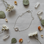 A delicate silver chain necklace with a sunburst moon charm, styled in a flat lay on a textured beige linen fabric surrounded by white flowers, green leaves, dried orange petals, and clear crystal points.