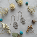 A pair of silver Hamsa hand drop earrings with intricate filigree cutouts, displayed on a textured light gray fabric background surrounded by dried flowers, green leaves, and small turquoise beads.