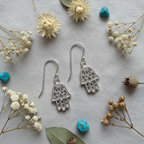 A pair of silver Hamsa hand drop earrings with intricate filigree cutouts, displayed on a textured light gray fabric background surrounded by dried flowers, green leaves, and small turquoise beads.