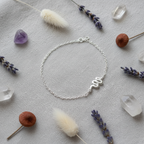 A delicate silver chain bracelet with a small wavy silver charm, displayed in a flat-lay arrangement on a textured light gray fabric surrounded by dried lavender, crystals, and fluffy white grass.