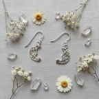 A pair of silver crescent moon drop earrings with intricate filigree cutouts, displayed on a textured light gray fabric background and surrounded by dried white flowers and clear crystal points.