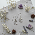 A pair of silver crescent moon and star dangle earrings arranged on a light gray textured fabric background, surrounded by dried white flowers, purple crystal clusters, lavender sprigs, and star anise.