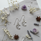 A pair of silver crescent moon and star dangle earrings arranged on a light gray textured fabric background, surrounded by dried white flowers, purple crystal clusters, lavender sprigs, and star anise.