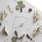 A delicate silver chain bracelet with a heart-shaped crystal pendant featuring a central turquoise stone, styled in a flat lay with white flowers, lavender sprigs, green leaves, and clear crystals on a light gray surface.