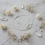 A delicate silver chain bracelet with a Hamsa hand pendant, styled in a flat-lay arrangement on a textured light gray linen fabric, surrounded by dried white flowers and clear crystal points.