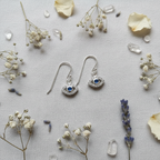 A pair of silver dangle earrings with evil eye charms, styled in a flat lay on a textured light gray fabric with dried baby's breath, lavender, petals, and clear crystals.