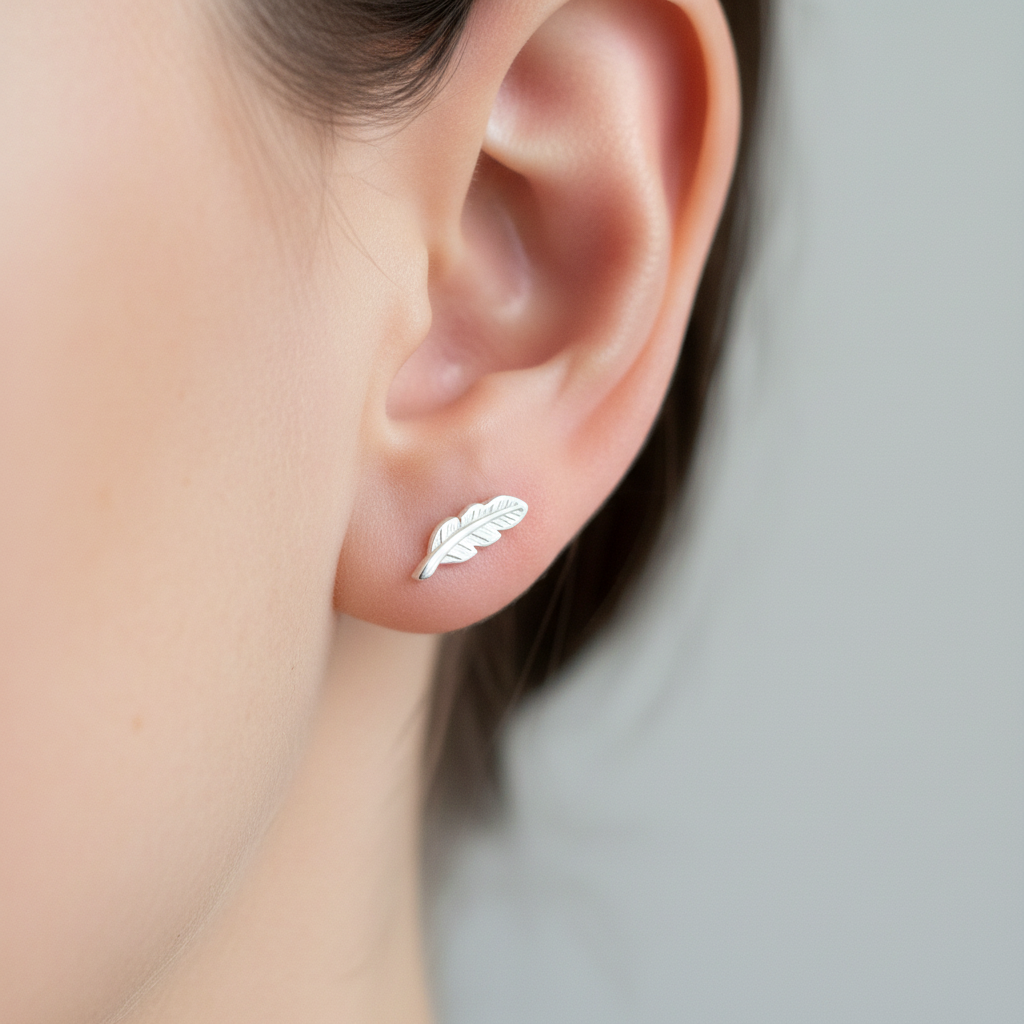 A close-up of a person's ear wearing a small silver leaf stud earring against a soft light gray background.