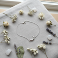 A delicate silver crescent moon bracelet with a small clear crystal accent, displayed on a light gray linen cloth surrounded by dried flowers, lavender sprigs, and white stones.