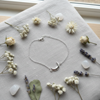 A delicate silver crescent moon bracelet with a small clear crystal accent, displayed on a light gray linen cloth surrounded by dried flowers, lavender sprigs, and white stones.