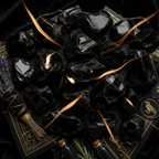 A styled flat-lay of glossy black polished stones arranged on a black tray with gold ornamental detailing, alongside a tassel, a small bottle, rosemary, and a wax seal with an owl emblem.