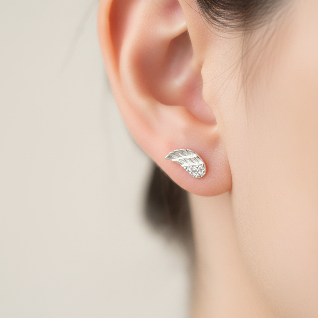 A close-up of a person's ear wearing a small silver leaf stud earring with tiny clear stones along the lower edge.