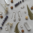 A pair of silver dangle earrings with a coiled, snake-like design, displayed on a textured light gray fabric background surrounded by dried botanicals, crystals, and star anise.