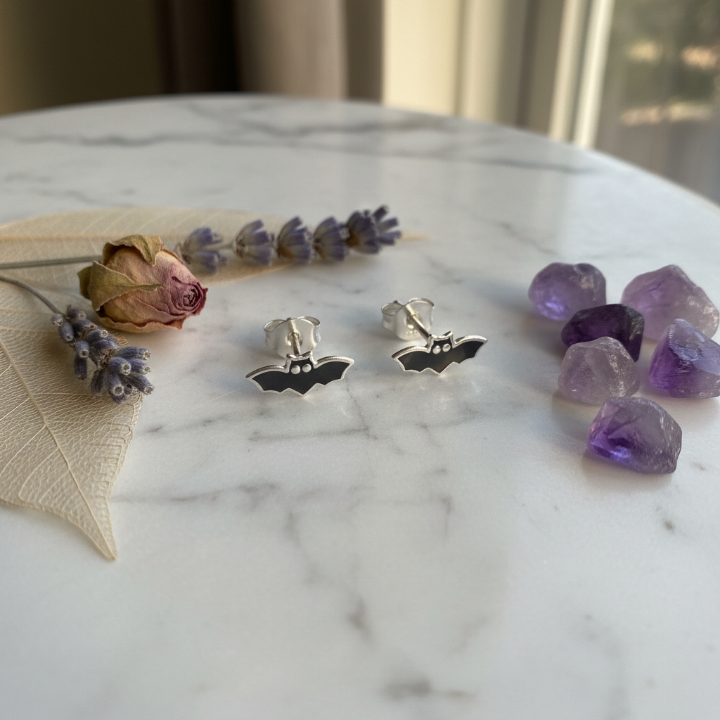 A pair of silver bat stud earrings displayed on a white marble surface, styled with dried lavender and purple crystals.