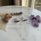 A pair of silver bat stud earrings displayed on a white marble surface, styled with dried lavender and purple crystals.